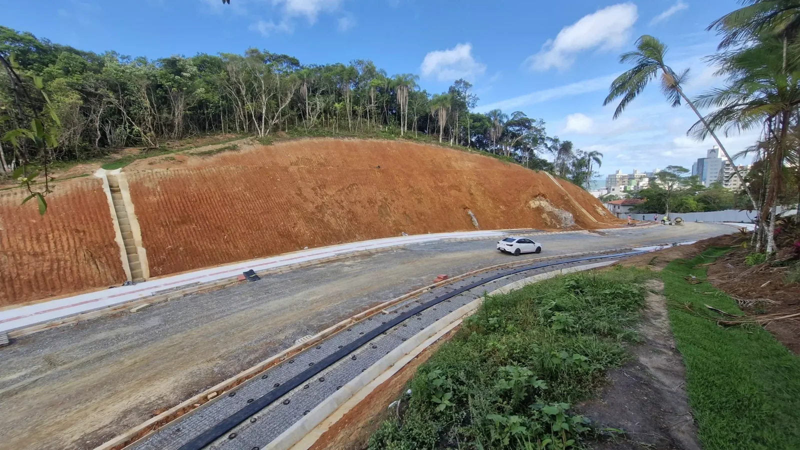 Trecho já executado do binário: pista nova de asfalto, meio-fio, drenagem e talude de contenção em terra vermelha, com prédios de Itajaí ao fundo